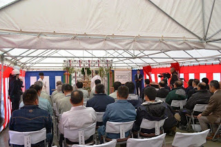 赤白の幕に囲まれたテントの中で、椅子に座った多くの関係者たちが前方の祭壇や演台を見ながら、建設に関する式典に参加している起工式の様子の写真