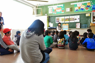 子どもたちが教室の床に向かい合って座り、前方に座った女性が紙しばいを使って話をしている様子の写真