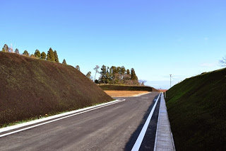 青空の下造成地へまっすぐ伸びている、両側を草で覆われた高台の間を通る、真新しいアスファルト舗装の道路の写真