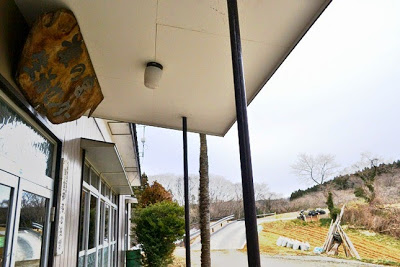 木製の看板が掛かっている建物の軒先から、外の道や整地された土地が見える写真