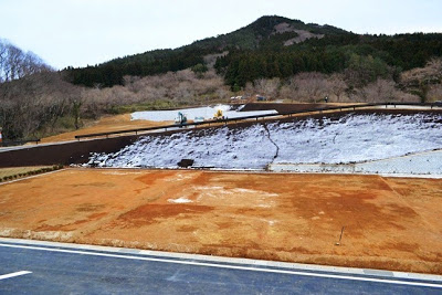 斜面に雪が残っている奥に重機が作業を行っている建設現場で、整地された土地が山を背に広がっている写真