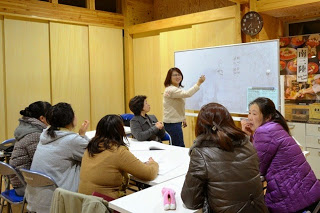 ホワイトボードの前に立つ白い服を着た女性講師が文字を指差しながら説明している様子を、受講者たちが座って熱心に話を聞いている写真