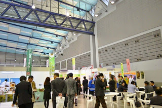 天井が高く光が差し込む広い室内で、多くの人々がブースを見学したり、白い丸テーブルの席に座って談話したりしている展示会場内部の様子を写した写真