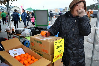 「こだわりみかん5キログラム2,000円」と書かれた黄色のポップが貼られた箱売りミカンが積まれているブースで、開いた箱に入っていたみかんを一個手に取る茶色のニット帽を被った笑顔の女性の写真
