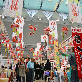 天井から飾りが吊るされ、カラフルなのぼりが並ぶ会場の通路を、多くの来場者が歩きながらブースを見て回っている様子の写真
