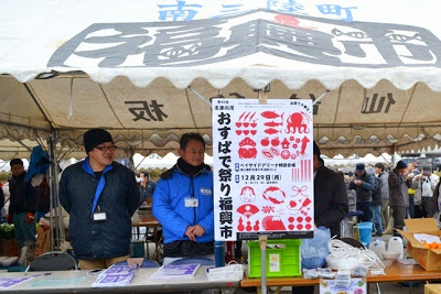 白いテントの下でチラシが置かれた長机の前に二人の男性スタッフが立っており、中央に「おすばで祭り福興市」と書かれたポスターが掲げられている南三陸町のイベント会場の写真