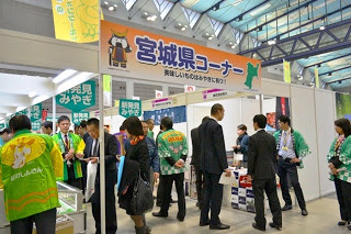 「宮城県コーナー」と書かれた看板の下で、来場者と出展者が商品の説明や試食を通して交流している様子を写した写真