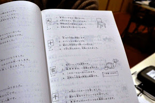 漢字の読み方やイラストが添えられた例文が書かれているページが開かれた、日本語の学習用テキストの写真