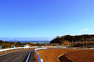 青い海と防波堤が見えている高台に、整備された新しい道路が大きくカーブしながら下っている風景の写真