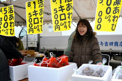 黄色の大きな値札が吊るされたテントの下で、笑顔の女性がアワビなどを発泡スチロール箱に入れて販売している魚介類販売ブースの写真