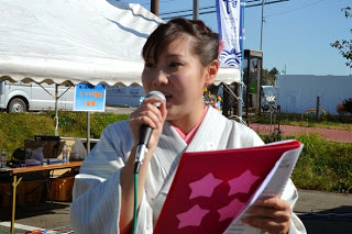 着物姿の女性がピンクのファイルを持ち、右手にマイクを持って進行を行っている様子をアップで撮影した写真