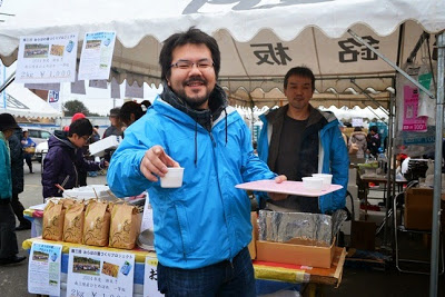青いジャンパーを着た男性が、にこやかに試食の紙コップを差し出している、地元特産品の試食ブースの写真
