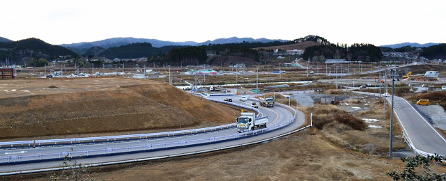 背景に造成中の広い土地と山々が見えるカーブした道路を車やトラックが連なって走行している写真
