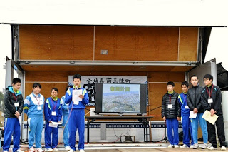 ステージの上に青いジャージを着た中学生たちが並び、一歩前に立っている男子生徒がマイクを持って発表をしている様子の写真