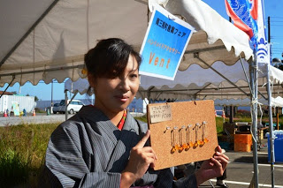 着物を着た女性が「Vent」と書かれた看板の下で、手作りのストラップの様な物を掲げて見せている写真