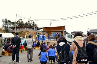 路上に設けられた仮設ステージの前でたくさんの人々が座り、中学生による発表を見ている様子の写真