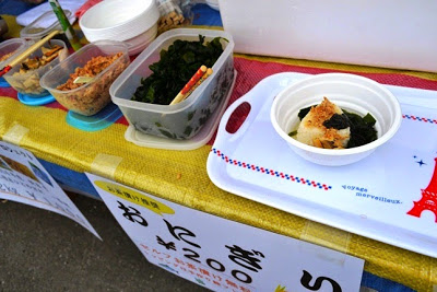 白いトレイの上に焼きおにぎりとワカメが盛られた料理が置かれており、周囲にはトッピングが並ぶ地元料理の屋台の写真