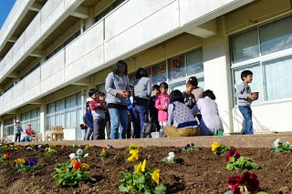 学校の建物の外で数人の子どもたちが集まり、花壇の前で植え付けの準備をしている様子の写真