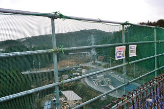 安全ネット越しに大型の重機や構造物が確認できる、建設中の橋の上から下の工事現場を見下ろす形で撮影されている、橋上からの工事風景の写真