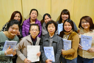 女性講師と参加者の9人の女性が日本語の教科書を手にして正面を向いて並び、教室の壁の前で記念撮影をしている写真