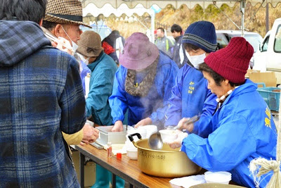 青いジャンパーとニット帽をかぶった女性たちが湯気を立てる大鍋で汁物をよそっており、列を作って並んでいる来場者に配っている写真