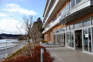 海沿いに建つ茶色と白の近代的な建物で、大きなガラス窓と自動ドアの入り口があり、歩道にはベンチと落葉した木々が並んでいる風景を捉えた写真