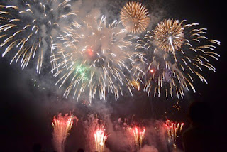 夜空いっぱいに広がる大輪の打ち上げ花火が赤や白、金色に輝き、煙の向こうから何発も連続して打ち上がっている盛大な花火大会の写真