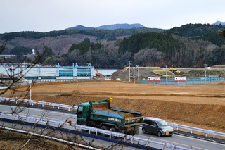 舗装道路をダンプカーと軽自動車が走り、その先に造成地と森、山々が見える写真