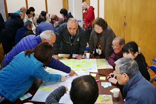 大勢の住民が机を囲み、地図の上にメモや付箋を貼りながら意見を出し合っている地域ワークショップの写真