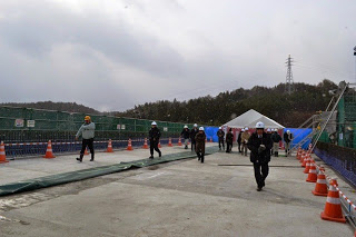 奥には白いテントが見える建設中の橋の上に仮設の歩道とコーンが並べられ、ヘルメットを被った関係者たちが歩いている、現場見学の写真