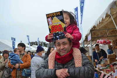 小さな女の子が父親に肩車されながら「おすばでまつり500円」と書かれたチラシを掲げ、楽しそうに笑っている親子連れの写真