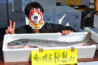 南三陸産の大きな秋鮭が発泡スチロールの箱に入って並べられ、傍にしゃがんだキャラクターのお面をつけた男性がピースをしている写真