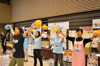 会場内のブースの前で、手に黄色や青色のポンポンを持った女性たちが元気に応援している写真