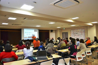 前方のスクリーンにはプレゼン資料が投影され、さまざまな年代の方がテーブルに着席しており、講師の話に耳を傾けている屋内の会議室の写真