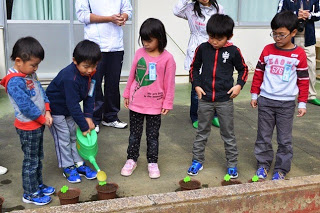 子どもたちが一列に並び、左から2番目の男の子が緑色のジョウロで植木鉢に水をあげている様子を、他の子どもたちが見ている写真