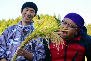 一人の男性が稲を手に持って笑顔を見せ、もう一人の男性がその稲を食べるふりをしてふざけている場面の写真