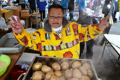 黄色い派手な柄のジャンパーを着た男性が、湯気の立つじゃがいもの大鍋の前で両手でピースサインをしている屋台の写真