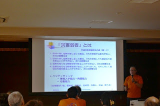 オレンジのTシャツを着用した関係者がマイクを持ちスクリーンの横に立ち、タイトル：「災害弱者」とはとかかれたセミナーのプレゼンテーションの内容がスクリーンに映し出されている写真