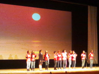 海の上に太陽があり、空が赤く染まる風景が映し出されている映像がステージのスクリーンに映し出され、その前に赤い服を着た複数の人々と制服を着た女子学生が横一列に並んで立ち、スタンドマイクに向かって歌っている写真