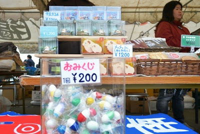 女性店員が奥に立っている販売ブースで、「おみくじ1個200円」と書かれたポップが貼られたテーブルの上にカプセル入りのおみくじが多数並び、後方に干支飾りなどが陳列されている写真