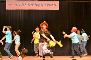 「テーマ：みんな生き活き！円結び」と書かれた横断幕が掲げられたステージで、オレンジ色のとんがり帽子を被った女性を中央に、色とりどりの服を着た参加者たちが笑顔で踊っている写真