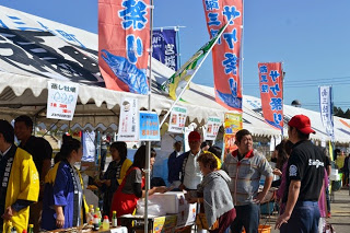 青空の下、色とりどりののぼり旗が立つテントの前で、多くの人が行列をつくりながら買い物をしている写真