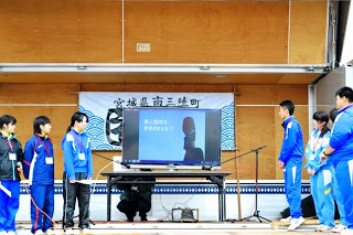 ステージ上の長机の上に設置された大型モニターに資料が映し出され、両脇に立つ中学生たちが説明を行っている様子の写真