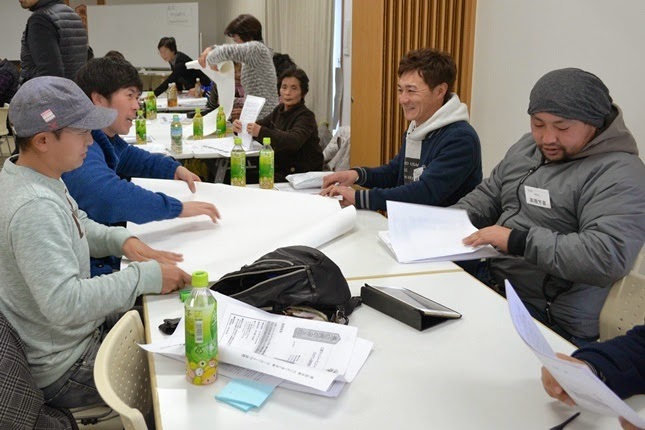 複数の男性が一つのテーブルを囲み、資料や模造紙を手にグループワークを行っている様子の写真