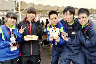 黒と青のジャージを着た中学生たちが、笑顔でカメラにピースサインをしながら食べ物やキャラクターの小物を見せている写真