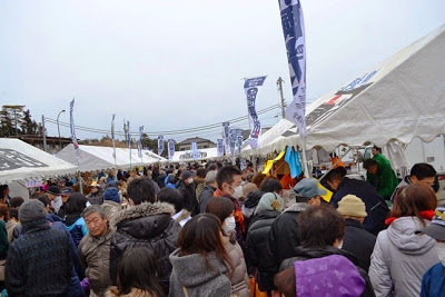 南三陸町の旗が立つ左右に並ぶ白いテントの間を多数の人々が歩いている、賑わいを見せる福興市の様子の写真