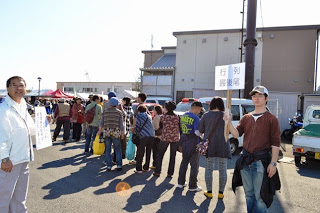 若い男性が「行列最後尾」と書かれた看板を高く掲げ、その前に多くの人々が列を作って並んでいる写真