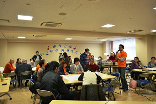 参加者がテーブルごとに分かれ、オレンジ色のTシャツを着た男性が前方でマイクを持って話している様子を参加者の人たちが耳を傾けている写真