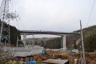 地上には重機と作業用資材が置かれており、森に囲まれた山あいにかかる高架橋が完成間近の状態の全景写真