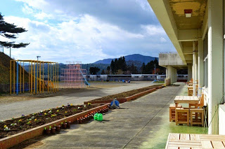 山々が見える穏やかな天気の空の下、学校の校舎沿いの花壇沿いに植木鉢が整然と並べられている、ジャングルジムなどの遊具が設置された校庭の写真